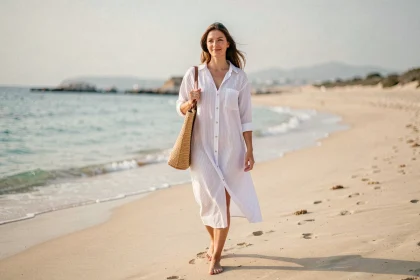 Femme portant une robe chemise blanche rayée marchant pieds nus sur une plage méditerranéenne avec un sac en paille, atmosphère décontractée et estivale