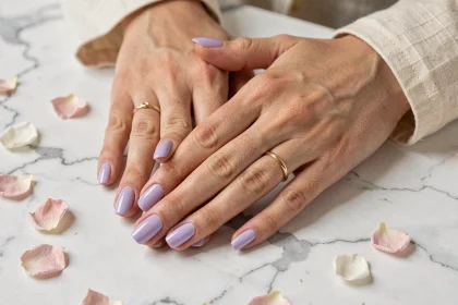 Mains de femme aux ongles vernis en pastel lilas posées sur du marbre blanc avec des pétales de fleurs printaniers